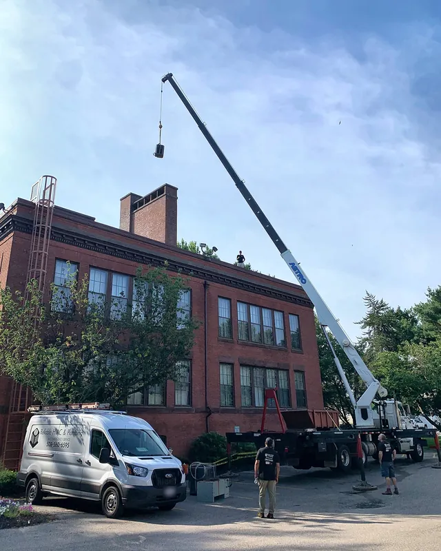 Crane lifting equipment onto the roof of a brick commercial building, with a Reliable HVAC van parked below