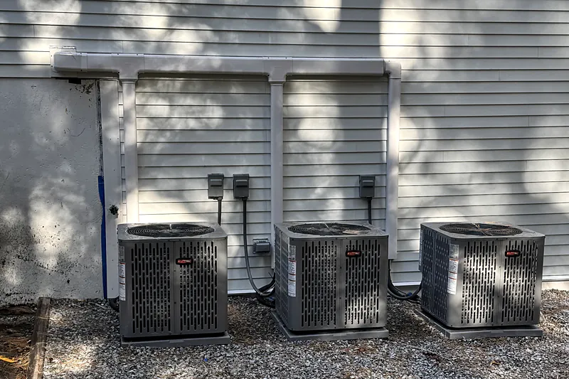 Three central air condensers on a gravel pad along the side of a house